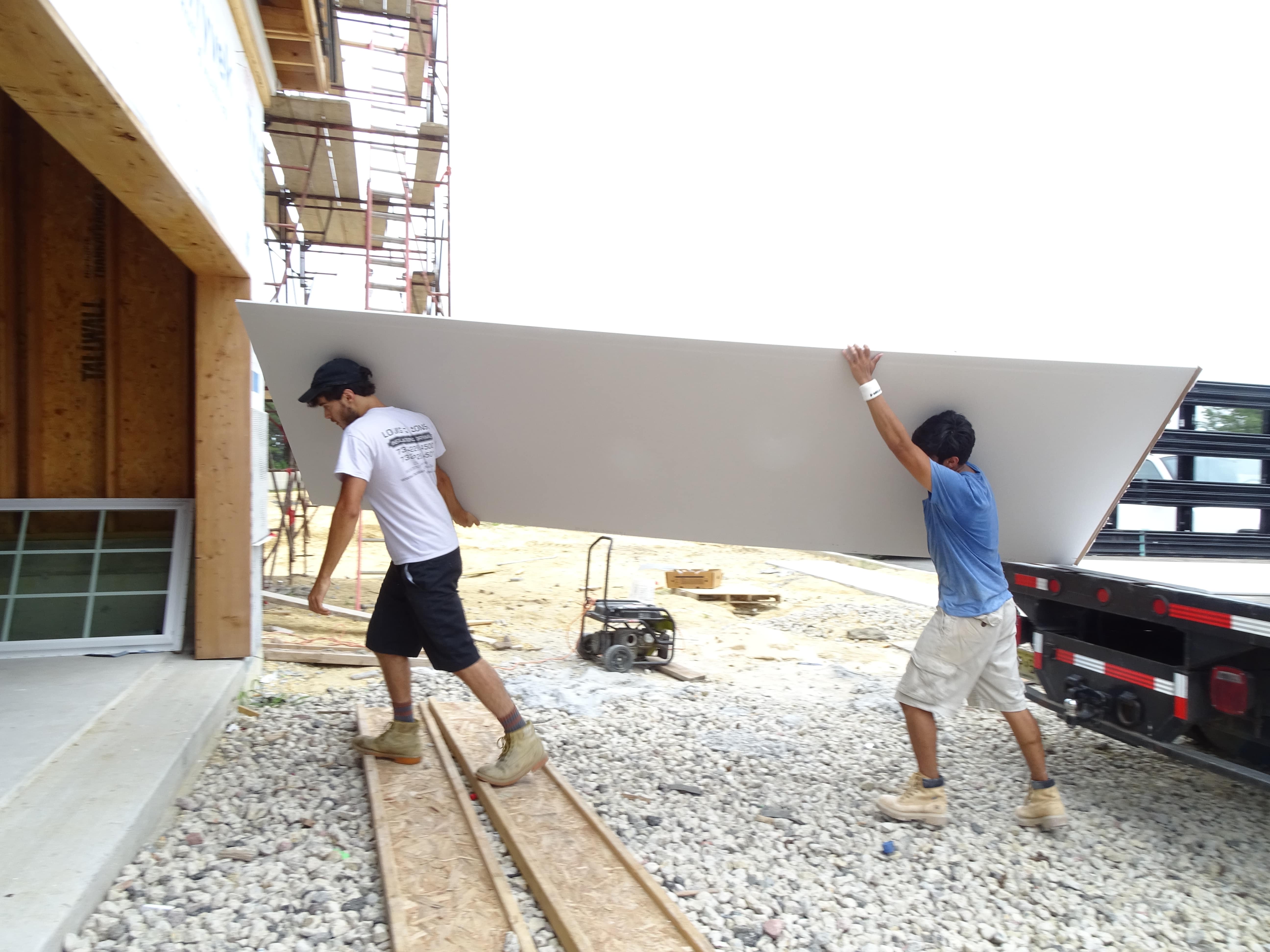 two workers carrying a sheet of drywall to the job location