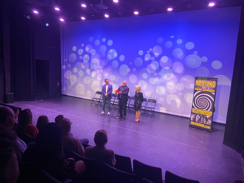 A stage with three people standing in front of a colorful backdrop, with chairs set up and a sign for Mr. Hypno on the side.